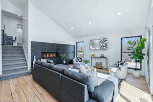 Living room with a tiled fireplace, high vaulted ceiling, light wood finished floors, stairway, and recessed lighting