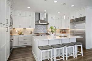 Kitchen with wall chimney exhaust hood, white cabinetry, built in appliances, a center island with sink, and a breakfast bar