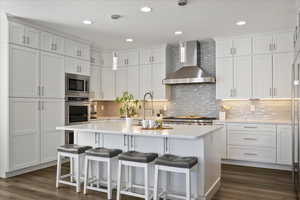 Kitchen with wall chimney range hood, white cabinets, a kitchen bar, hanging light fixtures, and dark wood-style flooring
