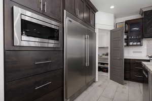 Kitchen featuring dark brown cabinets, built in appliances, glass insert cabinets, decorative backsplash, and exhaust hood