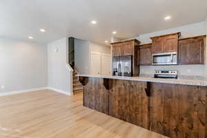 Kitchen featuring light wood-style flooring, appliances with stainless steel finishes, light stone countertops, a breakfast bar, and dark brown cabinets