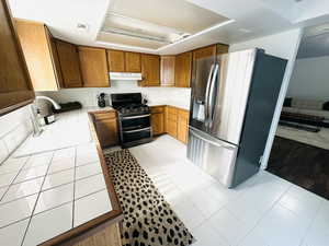 Kitchen with tile counters, brown cabinetry, stainless steel appliances, light tile patterned flooring, and under cabinet range hood