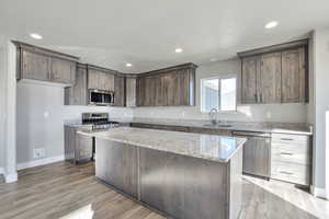 Kitchen featuring dark brown cabinetry, appliances with stainless steel finishes, a kitchen island, light stone counters, and light wood-type flooring