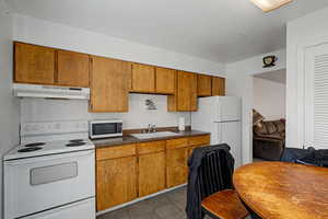 Kitchen featuring white appliances, dark countertops, brown cabinetry, under cabinet range hood, and dark tile patterned flooring