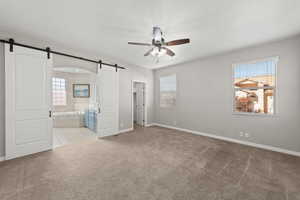 Unfurnished bedroom with a barn door, light colored carpet, ceiling fan, and ensuite bathroom
