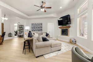 Living area with ceiling fan, a tiled fireplace, light wood-type flooring, and a raised ceiling