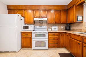 Kitchen featuring white appliances, brown cabinets, tasteful backsplash, under cabinet range hood, and light tile patterned floors