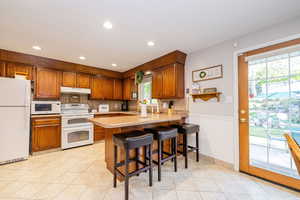 Kitchen with brown cabinets, white appliances, a breakfast bar, light countertops, and light tile patterned flooring