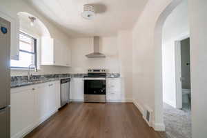Kitchen featuring arched walkways, stainless steel appliances, white cabinets, wall chimney exhaust hood, and ornamental molding