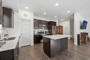 Kitchen featuring dark brown cabinets, stainless steel appliances, a kitchen island, dark wood finished floors, and light stone counters
