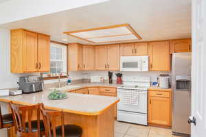 Kitchen with white appliances & stainless steel fridge, light countertops, a kitchen bar, light tile patterned flooring, and light oak cabinets.