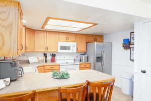 Kitchen with white appliances & stainless steel fridge, light countertops, a kitchen bar, light tile patterned flooring, and light oak cabinets.