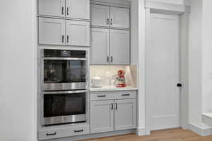 Kitchen featuring stainless steel double oven, gray cabinets, backsplash, light stone countertops, and light wood-type flooring