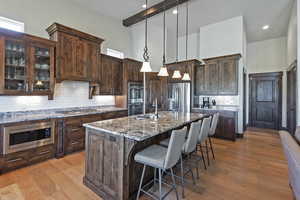 Kitchen with dark brown cabinets, backsplash, hanging light fixtures, light stone countertops, and light wood-style floors