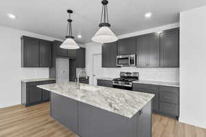 Kitchen featuring stainless steel appliances, light stone countertops, hanging light fixtures, an island with sink, and light wood-style floors