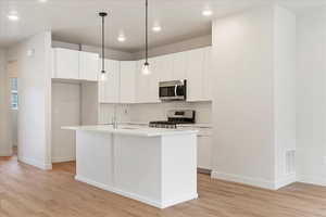 Kitchen featuring decorative light fixtures, white cabinetry, stainless steel appliances, a kitchen island with sink, and light wood-style flooring