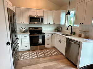 Kitchen featuring stainless steel appliances, vaulted ceiling, white cabinets, decorative light fixtures, and light wood-type flooring