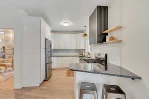 Kitchen with dark stone countertops, light wood finished floors, hanging light fixtures, open shelves, and a kitchen bar