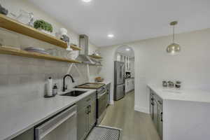 Kitchen with pendant lighting, appliances with stainless steel finishes, wall chimney range hood, light stone counters, and light wood-style flooring