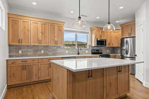 Kitchen with light wood finish cabinetry, stainless steel appliances, decorative light fixtures, light stone countertops, and a kitchen island