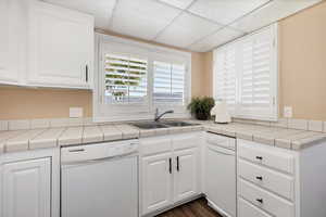 Kitchen with tile counters, white cabinetry, white dishwasher, a paneled ceiling, and dark wood-type flooring
