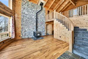 Unfurnished living room featuring high vaulted ceiling, a wood stove, a wooden ceiling with exposed beams, wood walls, and stairs