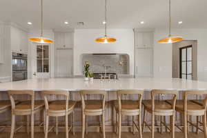 Kitchen featuring decorative backsplash, hanging light fixtures, stainless steel double oven, a large island with sink, and light stone countertops