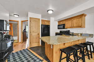 Kitchen with a peninsula, a kitchen bar, black appliances, light stone countertops, and wood finish cabinetry
