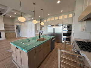 Kitchen with built in appliances, light wood finished floors, an island with sink, glass fronted cabinets, and range hood