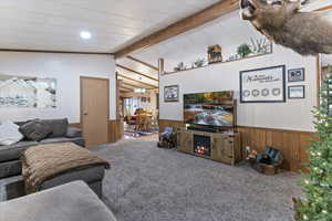 Carpeted living area featuring wooden walls, beam ceiling, a lit fireplace, and wainscoting