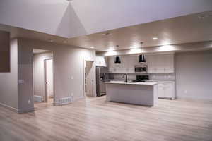 Kitchen with stainless steel appliances, a center island with sink, hanging light fixtures, light wood-style floors, and white cabinetry