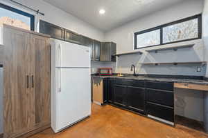 Kitchen with open shelves, finished concrete floors, freestanding refrigerator, dark cabinets, and a textured ceiling