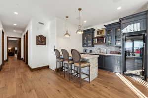 Indoor wet bar featuring glass fronted cabinets, hanging light fixtures, light stone countertops, decorative backsplash, and light wood-style flooring