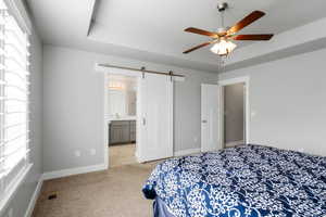 Bedroom with a barn door, a tray ceiling, carpet flooring, connected bathroom, and a ceiling fan
