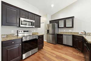Kitchen with stainless steel appliances, glass insert cabinets, light wood finished floors, light stone counters, and vaulted ceiling