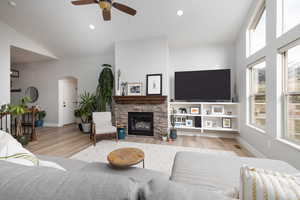 Living area featuring arched walkways, light wood-style floors, a stone fireplace, ceiling fan, and vaulted ceiling
