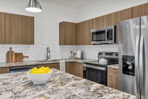 Kitchen with wood finish cabinets, stainless steel appliances, light stone counters, modern cabinets, and backsplash