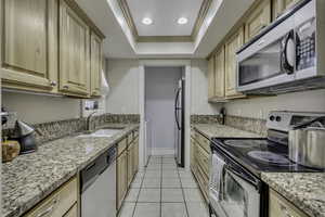 Kitchen featuring stainless steel appliances, ornamental molding, light stone countertops, a tray ceiling, and light tile patterned floors