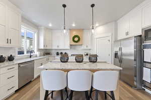 Kitchen with a breakfast bar, stainless steel appliances, white cabinets, light stone counters, and decorative light fixtures