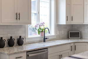 Kitchen with light stone counters, white cabinets, backsplash, stainless steel dishwasher, and healthy amount of natural light