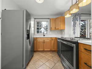 Kitchen featuring stainless steel appliances, light countertops, wood finish cabinetry, light tile patterned flooring, and hanging light fixtures