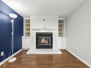Unfurnished living room featuring built in shelves, dark wood-style flooring, and a tiled fireplace