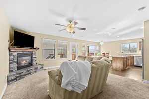 Living area with a stone fireplace, light carpet, and a ceiling fan