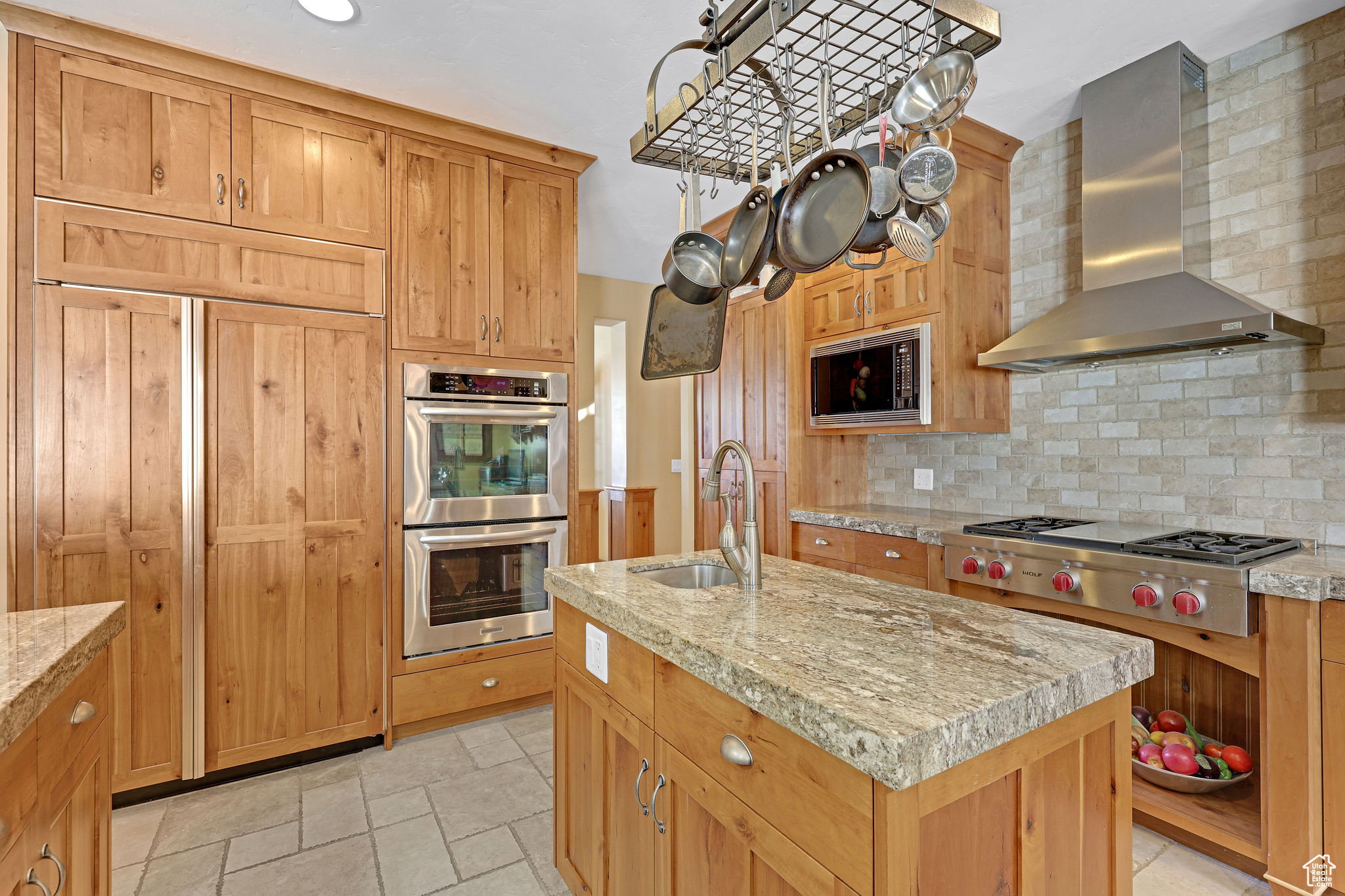 Kitchen with wall chimney range hood, built in appliances, a center island with sink, decorative backsplash, and stone tile floors