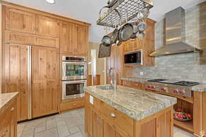 Kitchen with wall chimney range hood, built in appliances, a center island with sink, decorative backsplash, and stone tile floors