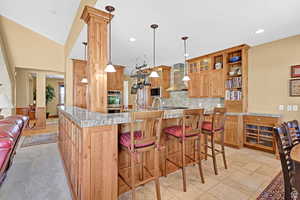 Kitchen featuring a kitchen breakfast bar, open shelves, glass insert cabinets, wall chimney range hood, and pendant lighting