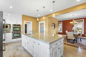 Kitchen with white cabinetry, stainless steel appliances, light stone counters, ornamental molding, and an island with sink
