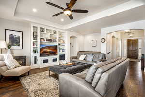 Living area featuring a raised ceiling, built in shelves, dark wood-style flooring, a ceiling fan, and a fireplace