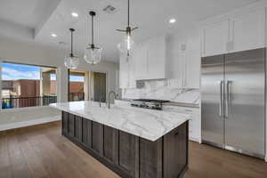 Kitchen with stainless steel appliances, dual tone cabinetry, dark wood-style flooring, light stone countertops, and decorative light fixtures