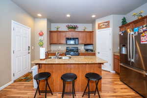 Kitchen featuring black appliances, wood finish cabinets, light stone countertops, light wood finished floors, and a kitchen island with sink